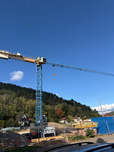 This image depicts a construction site located in Châtenois, France, on a clear, sunny day. A prominent, operational tower crane, primarily blue with a yellow jib section featuring the "G. Schreiber" logo, dominates the central frame, extending horizontally across the sky. Its hook assembly hangs idly.
Below the crane, the construction site is active, showing an excavated area with exposed soil and rebar for foundations. Piles of aggregates, stacked wooden formwork, and other construction materials are visible. An orange safety cone is positioned on the ground. Blue temporary fencing or large blue containers are situated on the right side of the site. In the far right background, a portion of a smaller, lighter-colored crane or utility pole is visible.
The background features a steep, densely forested hill with predominantly green trees, interspersed with patches of orange and brown autumn foliage. Several houses, some exhibiting traditional half-timbered architecture, are nestled at the base and on the lower slopes of the hill. The sky is bright blue with a few sparse white clouds. The very bottom of the frame shows the dark top of what appears to be a vehicle's hood. No individuals are visible in the scene.