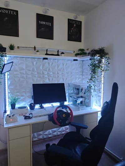 The image shows a gaming setup in a room. The setup includes a computer desk with a white desktop and drawers, a monitor, a white gaming PC case with visible components, a keyboard, a gaming wheel, and a white Xbox controller. Behind the desk is a wall with a 3D geometric pattern. LED strip lights are positioned along the edges of the wall. Above the desk are three framed posters with the word "VORTEX" and a shelf with decorative plants and a katana. A black gaming chair is positioned in front of the desk. The location, Stambolovo, Bulgaria, is not evident from the image.