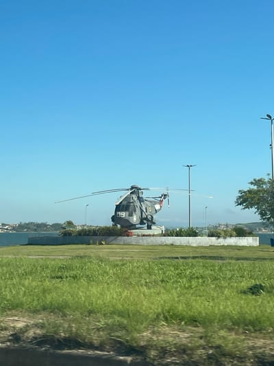 A gray helicopter with tail number 60 sits static on a platform with a concrete base. Adjacent to it is a grass area bordered by a concrete wall and lamp posts. Beyond this is a body of water. A clear blue sky fills the upper portion of the image.
