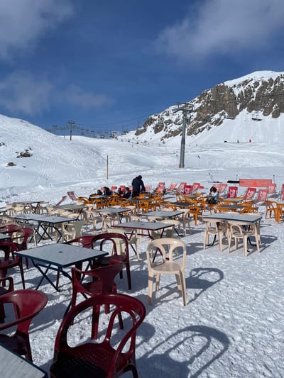 This image depicts an outdoor restaurant or café situated on a snow-covered mountain at a ski resort in Puy-Saint-André, France, under a clear blue sky with sparse clouds. In the foreground and mid-ground, numerous outdoor tables with light grey tops and various colored bases (dark blue, white, red) are arranged on the snow. Accompanying these are numerous plastic chairs in shades of dark red, orange, and beige. A section of the mid-ground features several red sun loungers, some displaying "Coca-Cola" branding.
Approximately ten to fifteen individuals are visible in the mid-ground, seated at tables or on loungers. One person in a dark jacket stands with their back to the viewer near a table, while another in a light blue jacket is seated at a different table. Several other individuals are less distinctly visible due to distance and focus.
In the background, snow-covered mountain slopes rise, displaying numerous ski tracks. A ski lift system, comprising multiple towers and attached chairs, traverses the slopes. Rocky outcrops are visible on the higher parts of the mountains to the right. The scene is brightly lit by natural sunlight, casting distinct, elongated shadows from the tables and chairs onto the snow. An orange banner or netting is visible in the mid-right background.