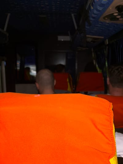 The image shows the interior of a bus with several passengers. In the foreground, a large orange cushion or seat cover dominates the lower half of the frame. Behind it, the back of a man's head is visible, with a shaved head. Further back, other passengers are seated, and their heads and shoulders are visible, with some wearing red and yellow clothing. The bus interior appears dimly lit, with some areas in shadow. The ceiling of the bus has a patterned blue design. The scene is consistent with the interior of a passenger bus, though no specific location within Kurnool, India, can be identified from the visual information alone.