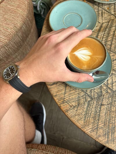 The image provides a close-up, first-person perspective of a scene inside an establishment in Kilmersdon, United Kingdom. The central focus is a human right hand, grasping a teal ceramic coffee cup containing a light brown beverage with white, heart-shaped latte art. The cup sits on a matching teal saucer, beside which a silver-toned spoon is partially visible. On the individual's left wrist, a silver-toned luxury sports watch with a black dial and a textured black rubber strap is displayed. The immediate surface is a round, distressed wooden table, characterized by numerous dark, linear scratch marks across its grain. Below the table, a bare human right thigh and a black athletic shoe with a white sole are partially visible. To the left, a section of a light-brown upholstered chair or bench, featuring a subtle striped pattern, is present. Blurred elements in the upper background include a green textile object and what appears to be an additional teal saucer, consistent with an indoor café or restaurant setting.