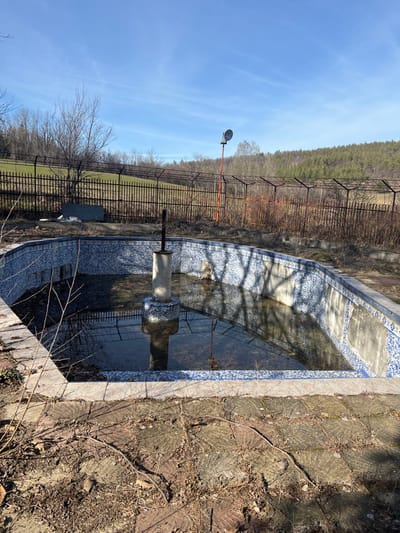 The image depicts an abandoned outdoor swimming pool or fountain located in Panichkovo, Bulgaria. The structure, roughly octagonal or rectangular with curved edges, is lined with blue and white patterned tiles, many of which are broken, missing, or discolored. It is partially filled with murky water containing organic debris such as leaves and branches. A central concrete pillar, also tiled, rises from the water, topped by a rusted, dark metal fixture. The immediate surrounding ground consists of discolored, circular-patterned paving stones covered with dry leaves, soil, and sparse vegetation. In the background, a rusty metal fence separates the pool area from a sloping landscape. To the left, a grassy field with bare deciduous trees is visible, while to the right, a densely forested hill with evergreen trees dominates. A tall, slender orange-red pole with a circular object (possibly a reflector or speaker) at its apex stands beyond the fence on the right. The sky above is clear blue with minimal cloud cover. No individuals or active subjects are present in the scene, which exhibits significant signs of neglect and disrepair.