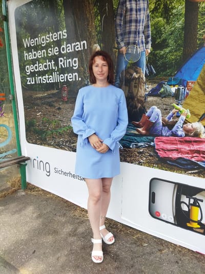 The image was taken in Monheim am Rhein, Germany, at a bus stop. A woman is standing in front of a large advertisement that reads, "Wenigstens haben sie daran gedacht, Ring zu installieren" (At least they thought about installing Ring). The advertisement depicts a camping scene with a family. The woman is wearing a light blue dress and white sandals. The bus stop shelter has a bench and is made of metal and glass. The advertisement appears to be on a bus stop shelter.