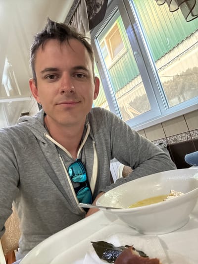 A man in a gray hoodie is sitting at a table with a bowl of soup and some food. He is taking a selfie. The man has short, dark hair and is wearing sunglasses clipped to his hoodie. Behind him, a window looks out onto a green corrugated metal building and a clear sky. The interior of the room appears to be a cafe or restaurant. The image was taken in the city of Yamantau, Russia.
