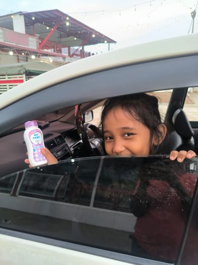A cheerful young girl, likely between 5-10 years old, is captured smiling broadly as she peeks out from the partially rolled-down window of a car. Her dark hair frames her face, and her hands rest on the car's window frame. She holds a white and purple bottle of "MY BABY baby powder" in her left hand, with the word "NEW" visible on the label.

Inside the car, parts of the dashboard, steering wheel, and a rearview mirror with an air freshener are visible. The exterior of the car appears to be a light beige or white. The background, seen through and above the car, reveals a multi-story building with a prominent red roof and red structural beams or railings. String lights with exposed bulbs are strung across the building's facade and in the sky, suggesting an evening or festive setting, though the overall sky appears overcast during the day, possibly late afternoon. Reflections on the car window show glimpses of the girl's red clothing and the external environment. The image conveys a happy, playful moment in Sukamulia, Indonesia.
