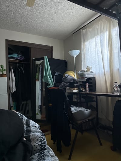 The image depicts a cluttered bedroom interior. A closet filled with clothing is visible on the left side of the frame. To the right, a desk is laden with various items, including a lamp, storage containers, a pink bottle, headphones, and a small white cat figurine. A chair with a light-colored cushion is positioned at the desk. A large window covered by sheer curtains is situated behind the desk. The flooring appears to be a yellow carpet. The scene is dimly lit, and the overall impression is one of disarray. The image was taken in Vancouver, United States of America.