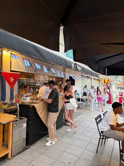 The image captures a lively street scene in Cambrils, Spain. Several people are seated at outdoor tables under a black awning, enjoying food and drinks. Cuban flags and bunting adorn the awning, suggesting a Cuban-themed establishment. Servers are seen attending to customers. In the background, pedestrians stroll along the sidewalk. The scene is well-lit, implying it is daytime or early evening. The foreground features bar equipment, indicating the location is a bar or restaurant.
