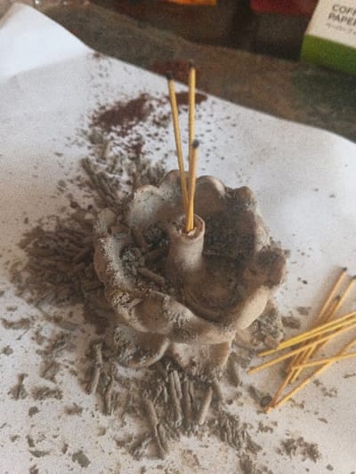 A top-down, slightly angled view captures an incense burning setup in Dogsenang Gorphoog, Bhutan, resting on a light-colored, possibly paper or cloth, surface. Central to the image is a weathered, light-brown, flower-shaped ceramic or clay incense burner. Two yellow incense sticks are vertically inserted into the central stem of the burner; their upper tips are blackened, indicating recent combustion. Surrounding the burner is a significant accumulation of fine grey ash and several darker, elongated, spent incense remnants. Towards the upper left, a scattering of darker reddish-brown granular material is intermixed with the ash. To the lower right, a small cluster of unlit, intact yellow incense sticks lies horizontally on the surface. The background, largely out of focus, reveals a dark, indistinct surface and the upper right corner of a green and white box with partial text "COFFEE PAPER". No individuals are present in the frame.