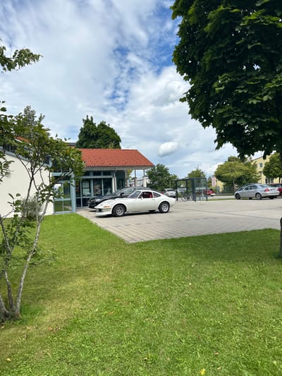 The image depicts an outdoor daytime scene at the Sparkasse Oberland - Geschäftsstelle Schongau-Mitte, located at Am Sportpark 1, 86956 Schongau, Germany.

The foreground features a manicured green lawn occupying the lower left and right portions of the frame. A deciduous tree with green foliage is partially visible in the lower left. In the midground, a light-colored building with a distinctive orange-red tiled roof and large glass windows forming an entrance is situated on the left. Adjacent to the building on the right is a grey paved parking lot constructed from interlocking pavers.

A white Opel GT classic sports car is parked prominently in the paved lot near the building entrance, oriented towards the left. Its red interior is visible. A dark-colored sedan is parked directly behind the white sports car, closer to the building. Further into the parking lot, a modern bus stop shelter with dark framing and transparent panels is visible. To the right, a silver sedan is parked. Additional vehicles and indistinct buildings are present in the far background. The sky overhead is partly cloudy, displaying white cumulus clouds against a blue background. No individuals are present in the scene.