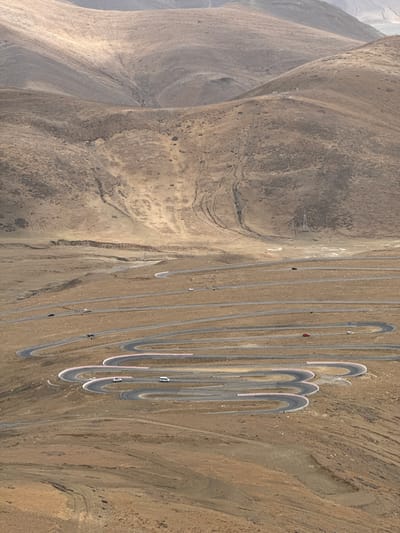 This aerial image shows a steep, arid mountainside with a winding road ascending it. The road is gray and features numerous hairpin turns, some marked with red and white barriers. Several vehicles are visible on the road. The terrain surrounding the road is predominantly brown and tan, typical of a desert or mountainous environment. The location is identified as Tashi Dzom town, China.