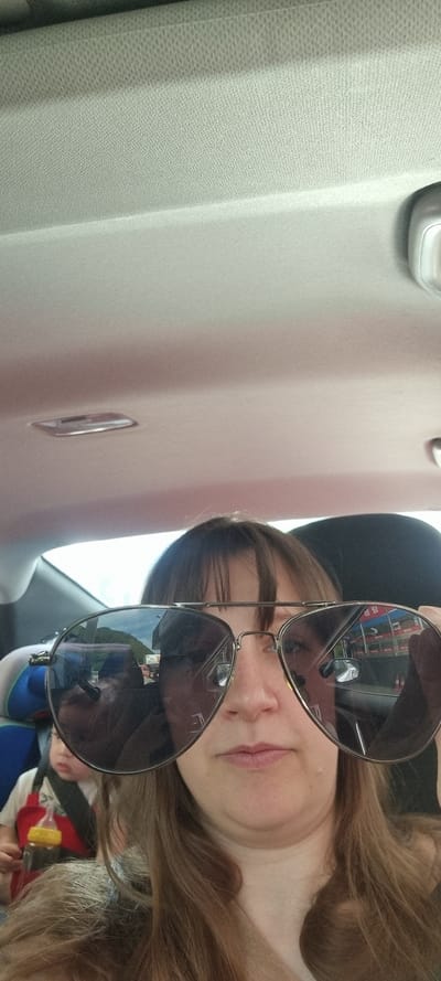 A woman is taking a selfie in a car in Yongpyeong-myeon, South Korea. She is wearing sunglasses that reflect the storefronts and buildings of the town. A child is visible in the background, possibly in a car seat. The interior of the car is also visible.