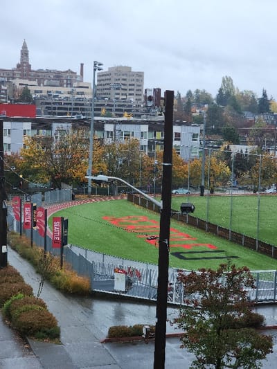 This image depicts an outdoor sports complex and its urban surroundings in Seattle, Washington, United States, specifically near Seattle University, under an overcast sky.

In the foreground, a wet concrete sidewalk runs diagonally from the lower left, transitioning into a wet asphalt parking area on the lower right. A dark utility pole stands vertically in the right foreground, partially obscuring a small deciduous tree with autumn-colored foliage.

The midground features a sports complex. A fence with vertical red banners displaying "SEATTLE U" and "REDHAWKS" separates the sidewalk from an artificial turf field. This field is surrounded by a reddish-brown running track. Large red letters spelling "REDHAWKS" and "SEATTLE U" are visible on the green turf. A dark piece of sports equipment rests on the field. Multiple tall poles with floodlights are positioned around the track.

The background consists of a dense urban environment with numerous buildings. Prominently featured is a tall, Gothic-style church steeple, identifiable as St. Ignatius Chapel of Seattle University. Other multi-story buildings, including one with an visible parking garage, are present. A hillside covered with trees, displaying autumn colors, and additional residential structures are visible behind these buildings.

The sky is uniformly grey and overcast, indicating a non-sunny day, consistent with the wet appearance of the pavement. No individuals are visible in the scene.