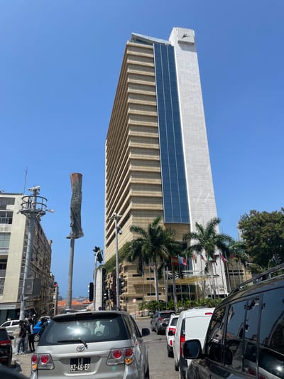 This vertical street view captures an urban scene in Luanda, Angola, under a bright, clear blue sky. The primary subject is a tall, modern building with a distinctive architectural design. The upper portion of the building features a smooth, light-colored facade and a prominent horizontal band of dark blue glass panels. Near the top left corner, a stylized logo, possibly depicting a bird or a letter, is visible. The lower sections of the building's facade are characterized by numerous horizontal, ribbed structures, creating a textured appearance.
In front of the building, several palm trees are visible, and a series of international flags are displayed on flagpoles, suggesting the location may be an embassy, a corporate headquarters, or a governmental institution. The street in the foreground and middle ground contains several vehicles. A silver SUV, identifiable by its Angolan license plate "LD 83-46-ER" (designating Luanda), is parked or moving on the right side of the frame. Other vehicles, including a white van and dark cars, are also present.
The scene depicts a typical bright, sunny day in a bustling city. Elements of street furniture, such as a pole with a traffic light and a tattered, possibly metallic structure, are visible in the middle ground, along with another pole featuring various antennas in the lower left. There are no clearly discernible people or specific activities beyond the general urban traffic. The visible license plate is the primary textual clue confirming the location.