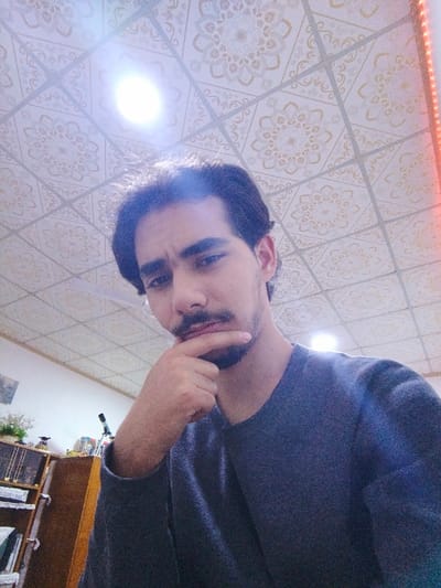 The image depicts a single male individual indoors within an unspecified building in Mosul, Iraq. The male, an adult with dark hair, a mustache, and a goatee, is centered and facing the camera with a direct gaze. His left hand is positioned with the index finger and thumb resting against his chin. He is wearing a dark grey long-sleeved shirt. The overhead ceiling consists of square, light-colored panels with a repetitive, circular ornamental pattern. A bright, circular overhead light fixture is visible in the upper-left quadrant of the ceiling, emitting a significant glow and lens flare. A vertical strip of red light is present along the top right edge of the frame. In the lower-left background, a partial view of a brown wooden shelving unit or bookshelf is visible, holding various indistinct objects, including a small green plant in a white pot and a black object resembling a telescope or camera on a tripod. The immediate wall behind the individual appears light-colored.