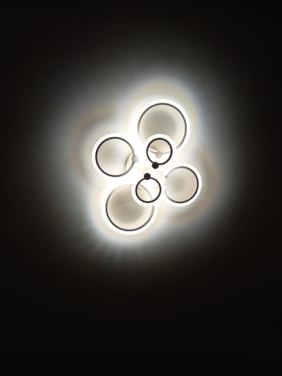 A modern, circular chandelier with multiple rings hangs in the center of a dimly lit room. The chandelier is illuminated, casting a bright white glow that fades into a dark, shadowy background. The rings vary in size and appear to be made of a metallic material with a matte black accent. Some of the rings have a translucent, textured edge where the light emanates. The overall impression is one of contemporary design and ambient lighting. The location in the city of Üshtobe, Kazakhstan, is not discernible from the image itself.
