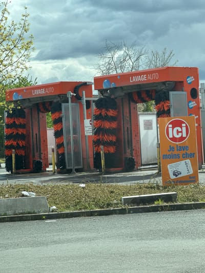 The image depicts an unoccupied automatic car wash facility located in Fleury-les-Aubrais, France, specifically identified as "FLEURY LAVAGE AUTO Commercial E.Leclerc Fleury Les Aubrais." Two identical, orange-colored car wash bays are centrally positioned under an overcast sky. Each bay features vertical and horizontal rotating brushes, primarily black with bright orange segments. The top section of each bay is labeled "LAVAGE AUTO" in white text, with additional labels for "BROSSE MOUSSE" (foam brush) and "LAVAGE CHASSIS" (chassis wash) visible on the rightmost bay.
In the foreground, to the right, a prominent orange vertical sign displays "ici Je lave moins cher" (Here I wash cheaper) in white text, with an additional note "Avec la carte d'abonnement" (With the subscription card) and contact details for the "FLEURY LAVAGE AUTO." A smaller white sign inside the left bay entrance states "INTERDIT AUX VÉHICULES ÉQUIPÉS DE GALERIES OU ACCESSOIRES" (Forbidden for vehicles equipped with roof racks or accessories).
The ground consists of asphalt pavement, separated from a strip of low, somewhat unkempt green and brown vegetation by a concrete curb and an intermittent, low concrete barrier. Several pieces of white litter, possibly plastic bags or wrappers, are scattered within this vegetation strip. Yellow bollards are visible at the entrance to the car wash bays. In the background, beyond the car wash structures, various trees with sparse or budding foliage are visible against the cloudy sky. No people or vehicles are present in the scene, and no actions or interactions are occurring.