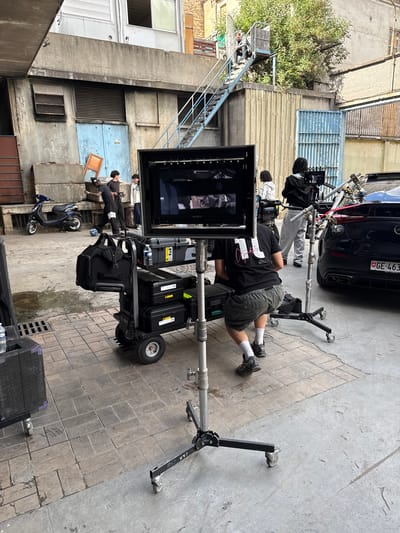 The image captures a film shoot in an urban alleyway, likely Ivry-sur-Seine, France, based on the car's license plate. A black car is partially visible in the background, alongside film equipment. A crew of people is present, including a person lying on the ground near the camera monitor, and others operating film equipment. The environment is a mix of concrete buildings with windows, doors, and a metal staircase. A blue scooter is also parked in the alley. The lighting suggests it's daytime.