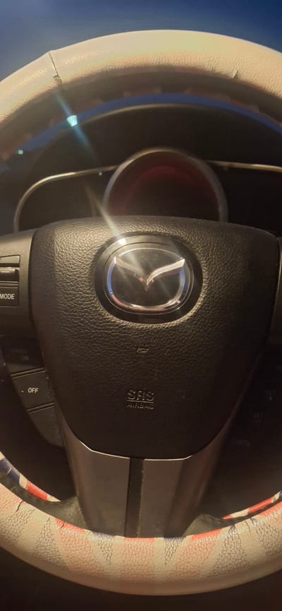 A close-up shot focuses on the steering wheel of a vehicle. The steering wheel is black and textured, with the silver Mazda logo prominently displayed in the center. On the left side of the steering wheel, there are several buttons, including one labeled "MODE" and another labeled "OFF". Below the Mazda logo, the text "SRS AIRBAG" is visible. The steering wheel is covered by a white steering wheel cover that features a pattern of red and blue stripes. The background is dark and appears to be the interior of a car, with a glimpse of the dashboard and possibly the speedometer visible. The image was captured in Sant'Egidio alla Vibrata, Italy.