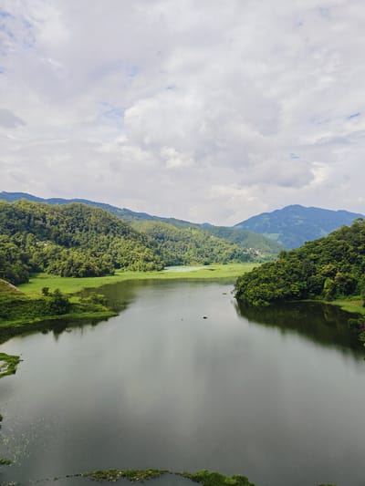A wide, still lake reflects the overcast sky and the lush green hills that surround it. The water is a dark, murky green, with patches of aquatic plants visible near the edges. The hills are densely forested with a variety of trees, creating a textured, undulating landscape. In the distance, softer, blue-hued mountains are visible, suggesting a vast, natural expanse. The sky is filled with a mixture of white and gray clouds, creating a diffused light that softens the scene. The image appears to be taken from a high vantage point, offering a panoramic view of the tranquil environment. The location is identified as the city of Lekhnath, Nepal, known for its numerous lakes and natural beauty.