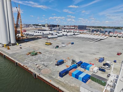 This aerial view captures a bustling industrial port in Esbjerg, Denmark, on a bright, partly cloudy day. The scene is dominated by the immense scale of infrastructure for wind turbine manufacturing and assembly. In the foreground, a large body of dark green water laps against a concrete and steel seawall.
The port itself is a vast expanse of grey gravel, meticulously organized for heavy industry. Prominently featured are several colossal, pale grey wind turbine tower sections, standing upright near the water's edge, hinting at their immense size. Towering cranes, one a bright yellow and orange behemoth, are actively engaged, suggesting ongoing construction or logistical operations.
Scattered across the yard are numerous other components of wind turbines, including long, curved blades arranged in rows and numerous cylindrical tower segments laid out horizontally. Various pieces of industrial equipment, such as forklifts, trucks, and mobile cranes, are visible, indicating a hive of activity. A collection of shipping containers in various shades of blue, orange, and brown are stacked and arranged, likely holding parts or serving as temporary storage.
Among the industrial elements, there are also smaller details like piles of gravel, stacks of materials, and what appear to be modular temporary buildings. A few vehicles, including a red truck, a white van, and a dark blue car, are parked or moving within the port. The background reveals a cityscape with buildings and more industrial structures, suggesting the port is integrated within a larger urban or industrial area.
The clear blue sky with scattered white clouds indicates fair weather, likely midday given the brightness and shadows. The overall impression is one of large-scale, organized industrial activity focused on renewable energy infrastructure. While no individuals are clearly discernible as subjects due to the distance, the presence of vehicles and equipment implies human presence and operation. No specific text is legible in the provided image.