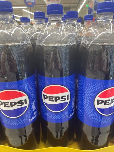 The image shows a close-up, slightly angled view of several plastic bottles of Pepsi soda. The bottles are arranged in a stack, with the labels clearly visible. The Pepsi logo, featuring the iconic red, white, and blue circle with the word "PEPSI" in white lettering, is prominent on each label. The blue portion of the label is decorated with a pattern of small white dots. Text on the labels indicates "1.5 Litre" and "Kola" (Turkish for cola). Some bottles also show nutritional information.
The setting appears to be a retail environment, possibly a supermarket or convenience store, given the presence of lighting fixtures visible in the background and what looks like a store sign with the word "Jeskosa" in the upper left corner. The bottles are likely on a shelf or in a display. The lighting suggests it is daytime, as might be expected in a store. There are no people visible in the image, and the focus is entirely on the product. The overall impression is one of a commercial display of beverages.