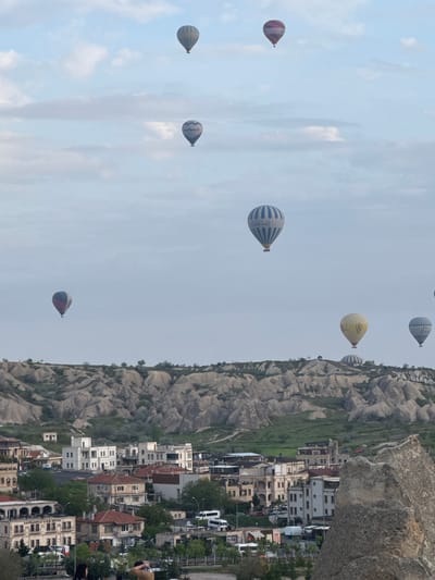 The image captures a scene in Göreme, Turkey, known for its unique landscape and hot air balloons. The sky is filled with hot air balloons of various colors and patterns, some with visible branding. Below, the terrain consists of distinctive rock formations and cave dwellings, typical of Cappadocia. Houses and buildings are nestled among the rocks, indicating a settlement area. The photograph is taken from an elevated position, providing a wide view of the valley.