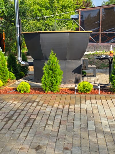 The image depicts an outdoor area, possibly a backyard or garden space, in Лужники Снт, Russia. The focal point is a large, black, conical cauldron-like structure with a wooden rim, which appears to be a large barbecue or cooking vessel. A stainless steel chimney extends vertically from the side of this structure. Surrounding the cauldron is a bed of red mulch, and several small, green evergreen bushes are planted around it. In the foreground, a paved walkway made of rectangular, multi-colored bricks is visible, appearing damp from recent rain. Behind the cauldron, a covered seating area or greenhouse-like structure with translucent panels and wooden framing is partially visible. A small metal table with a bottle of liquid and some tools or accessories rests to the right of the cauldron. The overall environment is lush and green, with dense foliage and trees in the background. There are no people visible in the image.
