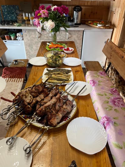 The image depicts a rustic dining table laden with food, suggesting a meal prepared at a dacha or a similar countryside dwelling. On the wooden table, a large platter of grilled meat skewers, known as shashlik, is prominently displayed. Accompanying the meat are dishes of small fried fish, a bowl of potato salad with herbs, and a platter of assorted fresh vegetables, including cherry tomatoes, cucumbers, carrots, and bell peppers. Two white, shell-shaped plates are positioned on the table, presumably for serving. In the background, a bouquet of vibrant peonies adds a touch of color. A white refrigerator is visible, along with kitchen cabinetry and a stove, indicating a functional kitchen area. The presence of a wooden bench covered with a floral blanket and stacked firewood in the corner suggests a cozy, informal setting, likely a dacha in Sovetskaya Gavan', Russia, a city known for its natural beauty and recreational opportunities, where such outdoor dining experiences are common.