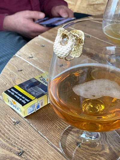 A table with two glasses, one filled with an amber liquid and some foam, a pack of yellow Camel cigarettes, and a gold-colored, woven-textured jewelry piece are visible. A person dressed in maroon and gray, and holding a phone, is partially shown on the side. The table surface appears to be made of wood planks, arranged in a pattern with metal fasteners.