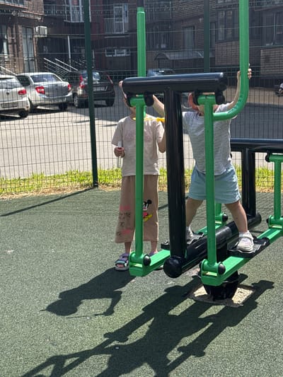 The image shows two children on a playground, likely in Vjazniki, Russia. One child is wearing a striped t-shirt and light blue shorts, with white sneakers. They are positioned on a green and black exercise machine, with their legs extended and hands gripping a green bar. The second child is wearing a light-colored t-shirt and beige pants with Mickey Mouse graphics. They are also on the exercise machine, in a lower position, with their hands on the black bars. The playground surface is a dark green, rubberized material. In the background, there is a chain-link fence, brick buildings, and parked cars, suggesting an urban or semi-urban setting. The presence of shadows indicates it is daytime, likely sunny. The overall mood seems to be one of outdoor recreation. Some text is visible on the second child's pants, including "LOVE TO" and "MR. RA".