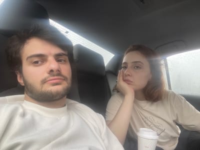 The image depicts two individuals inside a car. A woman with red hair is seated on the left side, holding a paper cup. A man with a beard is seated on the right. The car window shows raindrops, suggesting it is raining. The location is unidentified.