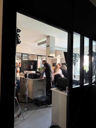 The image shows an interior scene, likely in a residential or commercial space, as viewed through a dark doorway or aperture.
Setting and Environment: The room inside has a modern aesthetic with white walls and gray flooring. A kitchen area is visible, featuring white cabinets, drawers, and a stainless steel ventilation system. A television screen with production equipment, cords, and camera equipment is set up in the kitchen. Daylight enters from windows in the distance.
People: Several people are present in the room, appearing to be involved in a film or video production. One individual with a tattoo is seen bending over, interacting with equipment on a counter. Two other individuals are lying on the floor. Another pair of people are in a kitchen setup behind the counter, and another obscured.
Objects and Equipment: Camera equipment, including tripods, cables, monitors, and a black equipment case, are present.
Actions and Interactions: The individuals appear to be setting up or operating film or video production equipment.
Location: While the request specifies Aubervilliers, France, this information is not visually apparent from the image alone.