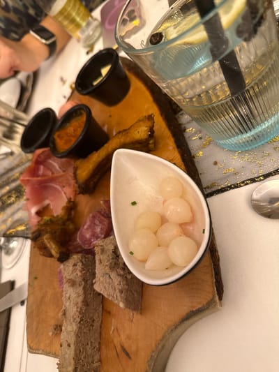 A wooden board displays various foods. Pickled onions sit in a white dish, while black ramekins likely contain condiments. Slices of bread and charcuterie, including salami and prosciutto, are arranged around a piece of fried bread. A pitcher of water is to the side. The overall impression is of a shared appetizer or charcuterie board.