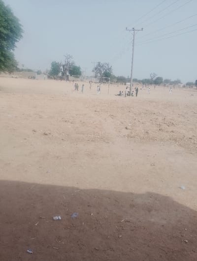 This image depicts an open, unpaved field serving as a community space in Kanwa, Nigeria. The ground is composed of dry, light brown earth, with minor debris including small white and grey objects in the foreground. A dark shadow extends across the lower section of the image from an unseen object to the left. In the mid-ground, a group of approximately 15-20 individuals, appearing to be children and young adults, are present. A primary group of 10-12 individuals is actively engaged in a game of soccer on a makeshift field, utilizing two rudimentary white goal frames. A soccer ball is visible near the players closest to the left goal. Another smaller group of approximately 5-7 individuals is gathered near the base of a prominent grey utility pole with multiple overhead power lines, situated to the right of the central playing area. In the background, distant, low-lying structures are discernible. Scattered green-leafed trees, including several large, mature specimens, punctuate the landscape in the mid-ground and background. The sky is a hazy light blue.