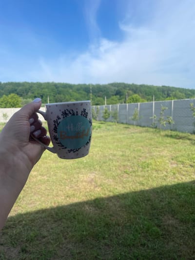 The image displays an outdoor scene captured on a clear, sunny day. A light-skinned hand, with fingernails painted a light, possibly lavender, shade, holds a white ceramic mug in the left mid-ground. The mug is adorned with a pattern of small pink dots and delicate black outlines of leaves and flowers. Centered on the mug is a teal-colored circular emblem containing the words "Hello Beautiful" written in a stylized, gold script.
The upper portion of the frame features a bright blue sky with sparse, wispy white clouds. Below the sky, a dense band of green foliage, indicative of a forest or wooded area, stretches across the horizon. Extending from the right side towards the center of the image is a long, grey pre-cast concrete fence, composed of uniform rectangular panels. Several small, young trees are planted at intervals in front of this fence.
The foreground and mid-ground are covered by a vibrant green grassy area, suggesting a lawn or field. A distinct diagonal shadow cuts across the bottom-left corner of the frame, indicating an unseen overhead structure to the immediate left. The scene portrays a tranquil, open-air environment, typical of a rural or suburban property. The specific location within the city of Petrivka, Ukraine, is not visually verifiable from the image content.
