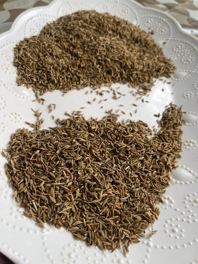 A high-angle, close-up photograph displays a collection of dried cumin seeds (Cuminum cyminum) distributed across a white ceramic plate. The plate features a decorative scalloped rim and an embossed pattern of small, raised circular dots. The cumin seeds are uniformly light to medium brown, elongated, and ridged. They are arranged in two primary clusters: a larger, less dense accumulation occupying the lower portion of the plate, and a smaller, denser pile positioned in the upper-left area. Scattered individual seeds bridge these two accumulations. The background appears as a blurred, patterned surface, indicating an indoor setting, likely a tabletop. No individuals, actions, or interactions are present within the frame. The location of this scene is Ksar-el-Kebir, Morocco.