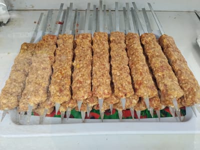 A close-up, high-angle shot shows ten raw ground meat kebabs on metal skewers, arranged in two rows on a white plastic tray. The kebabs are elongated and cylindrical, with a textured surface from the ground meat. They are lined up horizontally, with the points of the skewers visible at the bottom, resting on a colorful, patterned surface beneath the tray that features red and green elements. The background is a plain white surface, possibly a counter or shelf. The lighting is bright and even, casting no significant shadows. There are no people or animals visible in the image.