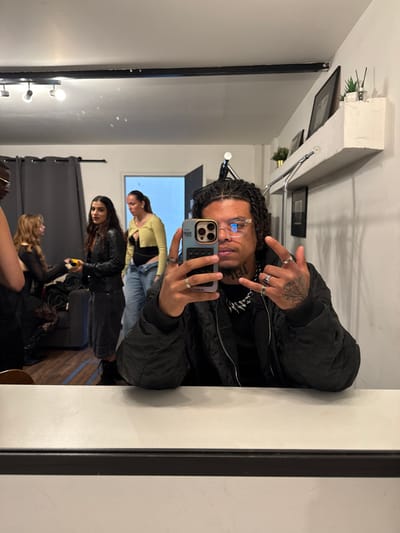 An indoor scene in Ivry-sur-Seine, France, is depicted through a mirror selfie. The primary subject, a male with dark, braided hair, clear-rimmed glasses, and visible facial and hand tattoos, is positioned in the lower right and center, reflected in the mirror. He wears a black quilted bomber jacket, a spiked necklace, and multiple rings. He holds a light-colored smartphone with a purple-hued case in his left hand, pointing it towards the mirror to capture the image. His right hand is raised, forming a "rock on" gesture.

The background, visible as a reflection, reveals a room with white walls and ceiling-mounted track lighting. On the left, a dark grey curtain or backdrop is present. Four other individuals are in the reflected space: a partially visible person on the far left, a woman with light brown hair in a dark top, a woman with long dark hair wearing a black leather jacket and holding a yellow object, and a woman in a yellow cardigan over a dark top and light blue jeans standing further back. A dark grey armchair is partially visible. An open light-colored doorway is seen in the rear-center. On the right wall, a white floating shelf holds framed art, a small plant, and a reed diffuser, with a metal rail and another framed picture below it.