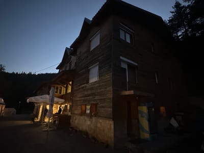 The image depicts a two-story wooden building with a dark, weathered facade, situated in an outdoor setting at dusk. The building has a sloped roof and multiple windows, some with closed shutters. In the foreground, there are two large white umbrellas, one of which is partially collapsed. Several people are visible, including two individuals standing near the umbrellas and one person seated at a wooden table. The ground in front of the building appears to be paved. A tall, thin lamp post is visible in the background to the left. The area is surrounded by trees, indicating a natural or semi-rural environment. The sky is a deep blue, suggesting twilight. There are also some coolers or vending machines near the entrance of the building. The building appears to be a commercial establishment, possibly a restaurant or lodge, given the outdoor seating and umbrellas. The overall impression is one of a quiet, possibly evening scene in a location that is not densely populated.