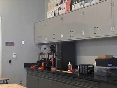 The image shows a gray-walled room with cabinets above a dark countertop. On the countertop are several 3D printers, spools of filament, and various supplies like glue, boxes, and tools. A black cabinet is situated between the printers and other supplies. The wall features a room number plaque "11-2350 Storage," a light switch, and a keycard reader. A wooden table with a stool sits in the foreground.