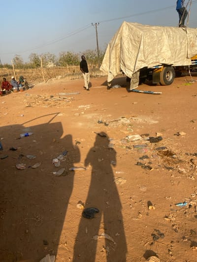 This image depicts an outdoor scene in the city of Tataura, Nigeria, characterized by an unpaved, reddish-brown dirt ground. The ground is extensively littered with discarded plastic bottles, bags, wrappers, and other debris. Long, distinct human shadows are cast from the lower-left foreground towards the center, indicating direct sunlight from a low angle. A partial shadow of a vehicle door is also visible in the lower-left.
In the midground, a large cargo truck is parked on the right. Its cargo area is covered by a light-colored tarp secured with ropes. A male individual wearing a blue shirt stands atop the tarp near the truck's rear, appearing to adjust or secure the covering. A blue and white striped object, possibly a pole, lies on the ground next to the truck's visible tire. To the left of the truck, another dark-skinned male, dressed in a black short-sleeved shirt and light-colored trousers, stands upright, hands near his pockets, observing the area.
Further to the left, a group of four to five individuals are seated or crouched on the ground beside a low concrete barrier. They are positioned near various containers, suggesting a market or vending activity. Dry, sparse vegetation, a dirt mound, and a rudimentary fence are visible in the far-left background. Overhead power lines and wooden utility poles are present in the background against a clear, light-colored sky.