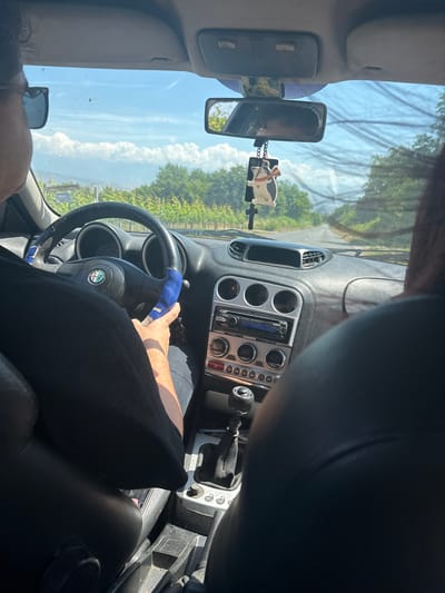 This image captures the interior of a car, viewed from the front passenger seat looking towards the driver's side and the windshield. The photo is rotated 90 degrees counter-clockwise. Bright daylight streams into the car, indicating it is daytime, likely midday or afternoon, under a clear blue sky with scattered white clouds. Outside the car, vibrant green trees are visible, suggesting a rural or natural landscape in Kolarovo, Bulgaria.
A person, presumably the driver, is seen with their right hand resting on the steering wheel. The steering wheel has a blue, somewhat worn cover and prominently features the "Alfa Romeo" logo in its center. The dark-colored dashboard includes a silver-toned center console housing a car radio, three circular gauges, air vents, and various control buttons. A manual gear stick with a black leather boot is visible to the right of the console.
A rearview mirror is centrally located, from which a distinctive "piggy" shaped car air freshener, black and white with a red scarf, hangs. A partial view of another person's head and hair is visible in the bottom left corner, indicating the presence of a passenger. The overall scene depicts a sunny journey through a scenic area. No specific text is clearly legible beyond the car brand logo.