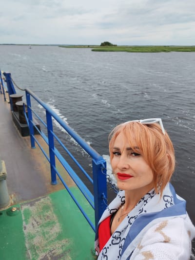 A woman with reddish-orange hair and red lipstick is taking a selfie on a ferry. She is wearing a red top, a white cardigan, and a patterned scarf. White sunglasses are perched on top of her head. The ferry is on a body of water, possibly a river or a bay, with a green and metallic deck and a blue railing visible. In the background, there is a flat landscape with green vegetation and a cloudy sky. The location is specified as Bereg, Russia.