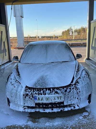 A black car, license plate AA 8023 YE registered in Ukraine, is covered in soap foam at a car wash. The car wash is an open-air structure with posts and signage, possibly marking the bays or services offered. The background shows a highway, a line of trees, a lamppost, and a clear sky. The photo appears to have been taken in the city of Shchaslyve, Ukraine, based on the license plate.