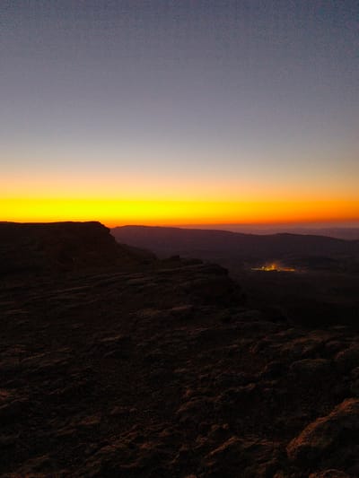 This image depicts a sunset or sunrise over the desert landscape surrounding Yeruham, Israel. The sky is a gradient of colors, transitioning from a deep blue at the top to a vibrant yellow and orange near the horizon. In the foreground, dark, rocky terrain suggests a high vantage point. In the middle ground, the silhouette of a cliff or plateau edge is visible. Further in the distance, a cluster of lights, likely representing the city of Yeruham, can be seen nestled among rolling hills, glowing against the darkening landscape. The overall scene conveys a sense of vastness and natural beauty.