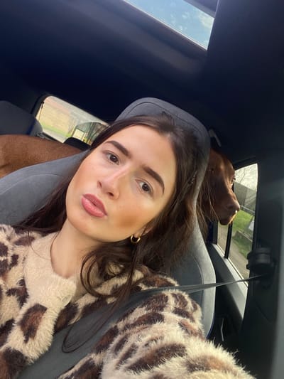 The image captures an interior car scene within a vehicle located in Broxbourne, United Kingdom. The primary subject is an adult female with long, dark brown hair, fair skin, prominent eyebrows, and light brown eyes. She is positioned in the front passenger seat, wearing a light-colored, fuzzy cardigan with a brown leopard print pattern, and her seatbelt is fastened across her chest. Her lips are slightly pursed, and she wears a light pinkish-brown lipstick, along with minimal facial makeup, including cheek contour and gold-colored hoop earrings. She is holding the camera for a selfie, angled slightly upwards towards her face.
Behind her, in the rear passenger compartment, a reddish-brown dog, possibly a Vizsla or similar breed, is partially visible. The dog's head is turned towards the right rear window, looking out, with its snout and part of its head illuminated by external light.
The vehicle's interior features dark blue or black upholstery on the ceiling, with a panoramic sunroof showing a bright, clear or lightly clouded blue sky. The car seats are grey fabric. Through the car windows, the exterior environment is visible, showing bare tree branches against a bright sky, and some green grass or low vegetation, indicative of a suburban or semi-rural setting. The time of day appears to be daylight.