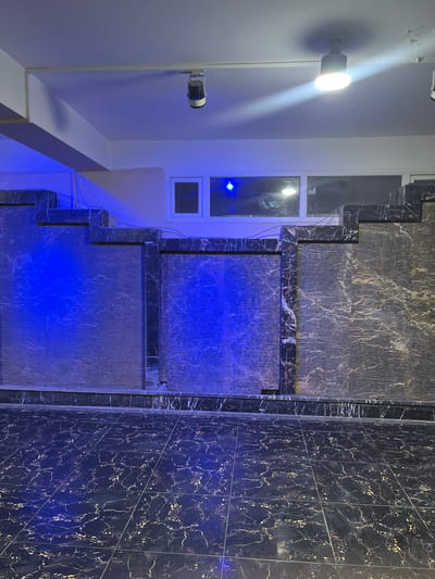 The image shows an indoor scene featuring a decorative wall with a water feature. The wall is clad in dark marble with intricate veining. The water feature appears to be made of textured panels, with blue lighting illuminating it. The floor is also tiled with dark marble, reflecting the blue light. Above the wall, there are windows and ceiling-mounted lights. One light fixture is clearly visible, emitting a bright white light, while another appears to be a blue spotlight. The overall impression is of a modern, stylish interior space, possibly a hotel lobby or a restaurant. The specific location in Kabul, Afghanistan, cannot be determined from the image alone.