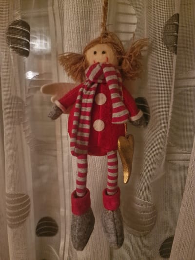 A handmade plush doll, resembling an angel, is suspended in front of a sheer, patterned curtain. The doll has light brown yarn hair styled in pigtails, and a simple fabric face with two black bead eyes and a red stitched mouth. It wears a red coat with three white buttons, a striped scarf in red and grey, and striped stockings in the same pattern. Its feet are encased in grey felt boots, and it has a single light-colored wing attached to its back. A golden, heart-shaped ornament hangs from the doll's right side. The curtain behind the doll is light-colored and features circular, semi-transparent patterns. The image was taken in Neuwied, Germany.