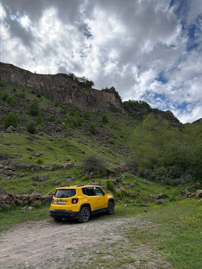 A yellow Jeep Renegade is parked on a gravel road in a mountainous, grassy landscape. The car is facing away from the camera, positioned on the left side of the frame. Behind the car, the terrain slopes upward, covered in green grass and scattered rocks. Further up the slope, a steep, rocky cliff face rises, partially covered with sparse vegetation. Trees are visible on the slopes and along the top of the cliff. The sky above is filled with dramatic, grey and white clouds, with patches of blue sky visible. The license plate of the Jeep reads "FG-661-FF". The overall scene depicts a remote, natural environment.